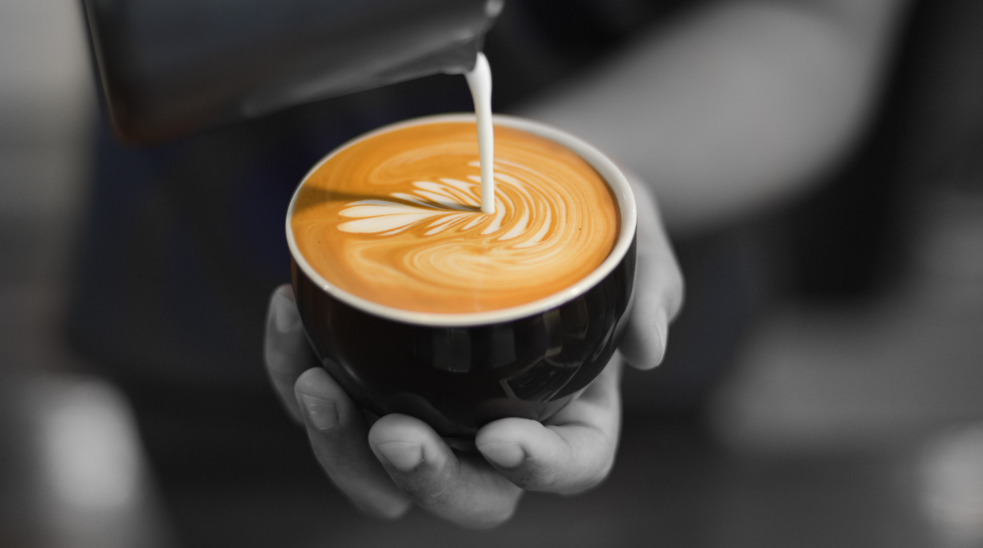 A barista pouring steamed milk from a steel jug into a cup of espresso, forming a leaf-shaped latte art pattern
